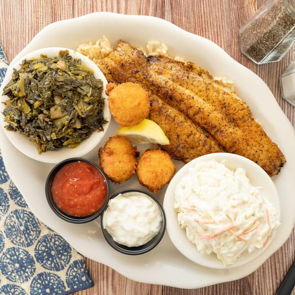 PoFolks' fried fish platter with collard greens, hush puppies, coleslaw, and sauces on a white plate.