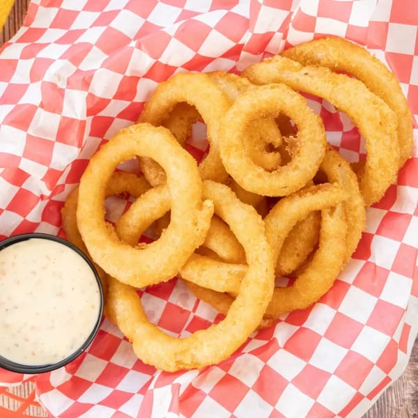 Golden-brown onion rings in a red and white checkered basket with a side of dipping sauce.