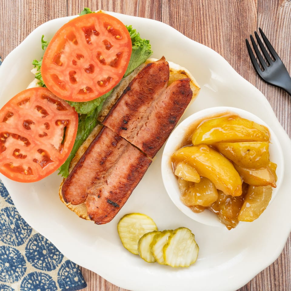 PoFolks' grilled hot dog with lettuce, tomato, a side of peaches, and pickles on a white plate.
